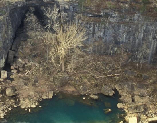 Photo of cliff face, talus, Jacks Fork River, and leafless injured sycamore trees